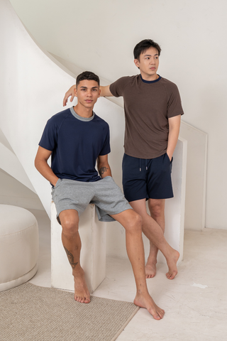 Two men wearing casual t-shirts and Olive Basics Noah Multi-Pocket Stretch Shorts pose indoors on a white staircase. One sits with hands in his pockets; the other stands barefoot, leaning with hands in his pockets.