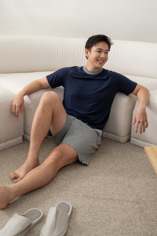 A man in a navy T-shirt and Olive Basics Noah Multi-Pocket Stretch Shorts sits on the floor, leaning against a white sofa. Slippers are on the carpet nearby.