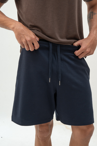 A person wearing a brown t-shirt and Olive Basics Noah Multi-Pocket Stretch Shorts in navy stands against a plain white background, hands adjusting the waistband.