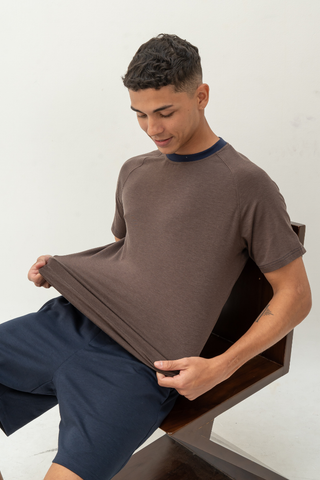 A young man sitting on a wooden chair pulls the front of his Noah Lightweight Multi-functional Tee by Olive Basics outward while looking down at it. He is also wearing navy blue shorts.