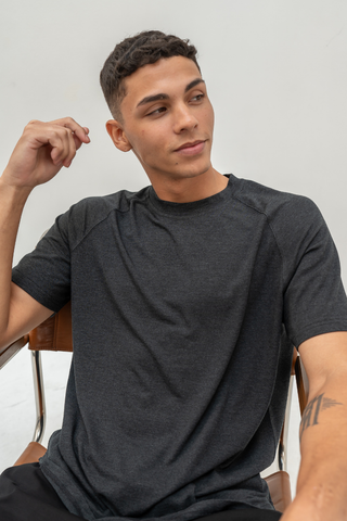A person with short curly hair sits on a chair, wearing the Olive Basics Noah Lightweight Multi-functional Tee in Powder Black, looking to the side against a plain light background.