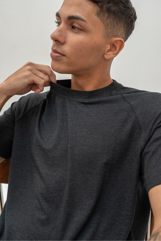 Wearing the Olive Basics Noah Lightweight Multi-functional Tee in dark gray, a person sits on a chair, gazing to the side and gently pulling at the shirt’s collar with one hand.