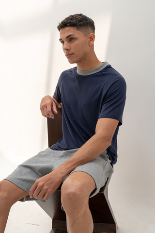 A young man sits on a chair in a brightly lit room with a white wall background, wearing the Olive Basics Noah Lightweight Multi-functional Tee in navy paired with gray shorts.