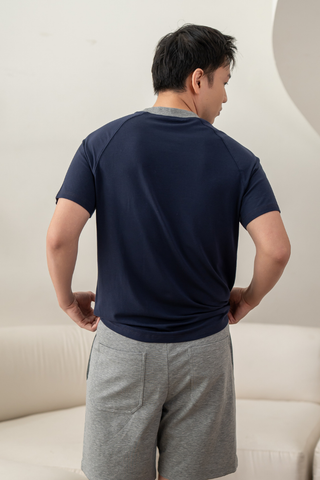 A man stands indoors with his back facing the camera, wearing the Olive Basics Noah Lightweight Multi-functional Tee in navy and gray shorts, with his hands resting on his hips.