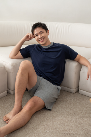 A man in the Olive Basics Noah Lightweight Multi-functional Tee in navy and gray shorts sits on the floor next to a white sofa, smiling at the camera.