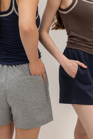 Two people, facing away from the camera against a plain light background, wear tank tops paired with Olive Basics Charissa Gentle Waist Pocket Shorts, each with their hands in their pockets.