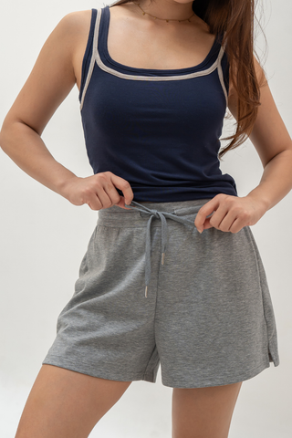 A woman stands against a plain background with her hands on the waistband of her Charissa Gentle Waist Pocket Shorts by Olive Basics, paired with a navy tank top.