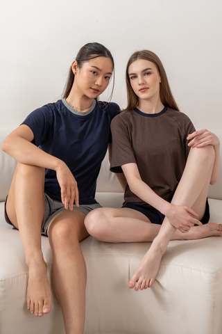 Two women sit side by side on a couch, wearing casual lounge sets by Olive Basics that feature the Chloe Relaxed Croppable Tee and matching shorts in soft TENCEL™ Wool Spandex, looking at the camera with neutral expressions.