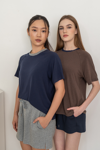Two women stand side by side against a plain wall, both wearing the Olive Basics Chloe Relaxed Croppable Tee and comfy lounge set shorts with their hands in their pockets, looking directly at the camera.