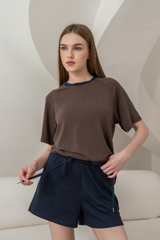 A woman stands indoors wearing a brown short-sleeve t-shirt and Olive Basics Charissa Gentle Waist Pocket Shorts in Navy, holding the drawstring at the gentle waistband of her shorts with a neutral expression.