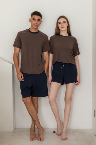Two people stand indoors against a white wall, both wearing brown T-shirts and Olive Basics Noah Multi-Pocket Stretch Shorts in navy. They are barefoot and looking at the camera with neutral expressions.