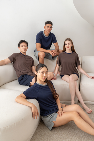 Four people in casual shirts and shorts, including the Olive Basics Noah Lightweight Multi-functional Tee crafted from a TENCEL™ wool blend, sit on a white sectional sofa in a bright room, facing the camera with neutral expressions.