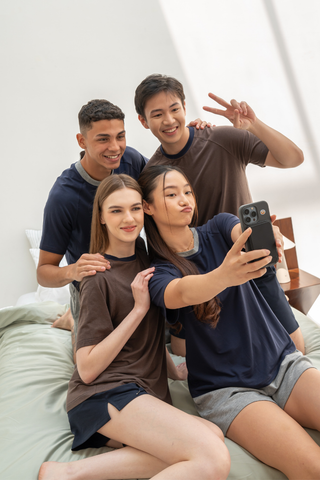 Four young adults sit on a bed, posing and smiling for a selfie; one person holds the phone while another flashes a peace sign, all comfortably dressed in cozy loungewear featuring the Olive Basics Chloe Relaxed Croppable Tee crafted from TENCEL™ Wool Spandex.