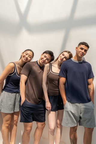 Four people stand close together, dressed in casual shorts and t-shirts—one wearing the stylish Chloe Braless Reversible Tank by Olive Basics—smiling and posing against a white wall in natural sunlight.