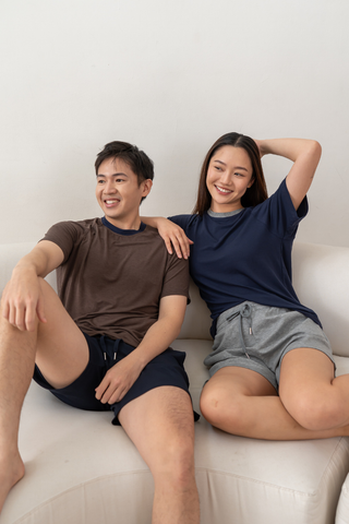 Two people sit on a white couch, both smiling. The person on the left wears a brown shirt and Olive Basics Charissa Gentle Waist Pocket Shorts in navy; the person on the right wears a navy shirt and gray TENCEL™ Modal shorts.