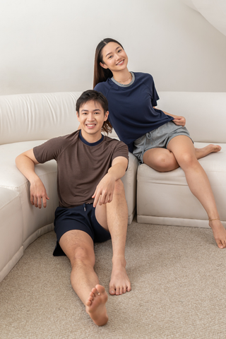 Two people are sitting on a white couch over a carpeted floor, both dressed in casual t-shirts—such as the Chloe Relaxed Croppable Tee from Olive Basics—and shorts, smiling at the camera in a bright indoor space. Their cozy lounge set from Olive Basics creates a comfortable and inviting atmosphere.