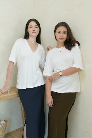 Two women stand indoors against a light wall, both wearing Olive Basics' Tabitha Reversible Multi-way Tee in white and long skirts—one navy, one olive with an adjustable waist—their expressions neutral.