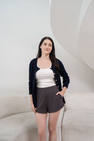 A black hair & fair skin women wearing a white fitted tank top layered with a navy button-up cardigan and Olive Basics Rebecca Expandable shorts, standing indoors against a minimal neutral background.