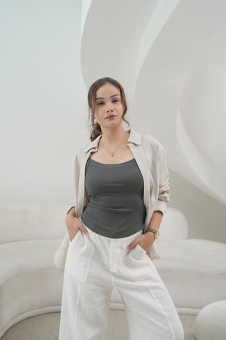 A woman stands indoors against a modern white curved backdrop, wearing the Olive Basics Diana V-neck Reversible Padded Tank in gray with white pants and an open light shirt, her hands resting in her pockets.