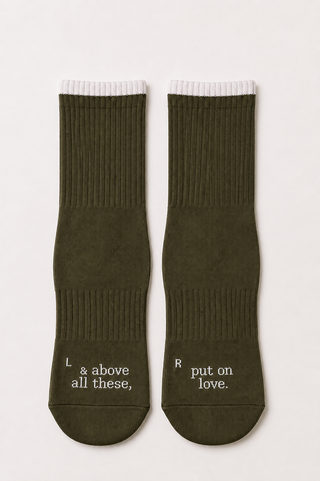 Cloudy Grip Socks by Olive Basics are a pair of khaki green grip socks with white cuffs; the left sock features the phrase “& above all these,” while the right sock reads “put on love.” Crafted from soft organic cotton, these pilates socks offer both comfort and thoughtful style.