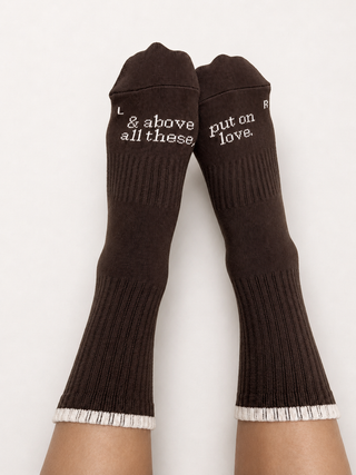 A pair of feet wearing Olive Basics Cloudy Grip Socks in brown, featuring white text that reads “L: & above all these, R: put on love” and white trim at the toes. These cozy socks are perfect for pilates or lounging in comfort and style.