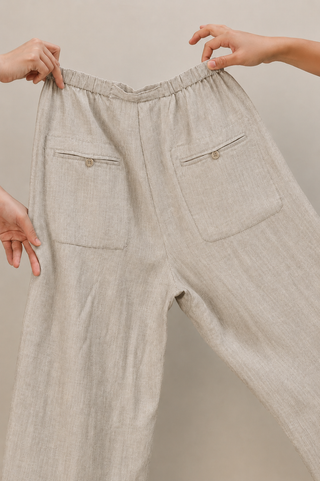 Two hands hold up the Olive Basics Bella Multi-Pocket Drawstring Pants in oatmeal, displaying the elastic waistband and two buttoned back pockets.