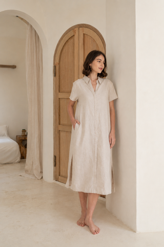 A woman stands barefoot indoors by a wooden arched door, wearing the Olive Basics Marie Short Sleeve Midi Dress in Heather Oatmeal.