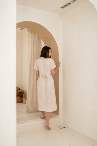 A woman wearing the Olive Basics Marie Short Sleeve Midi Dress stands barefoot, facing away, with her hand resting on a curved archway in a minimalist, neutral-toned room.