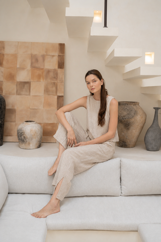 A woman in the Olive Basics Leigh-Anne Reversible Button Top in Heather Oatmeal with breastfeeding access sits barefoot on a light gray sofa, surrounded by large ceramic vases and modern neutral decor with floating stairs behind her.
