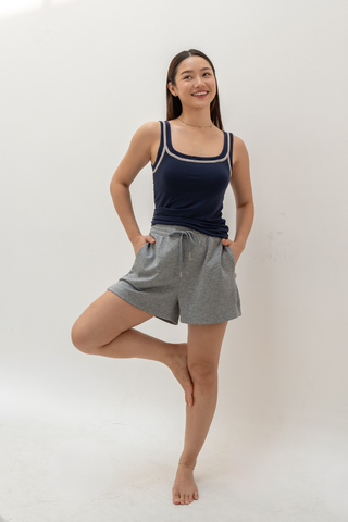 A woman stands on one leg in a yoga pose, wearing a navy tank top and Olive Basics Charissa Gentle Waist Pocket Shorts in heather grey, set against a neutral background.