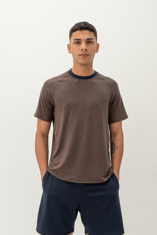 A person stands against a plain light background wearing the Olive Basics Noah Lightweight Multi-functional Tee in chocolate and dark shorts, with hands in pockets.