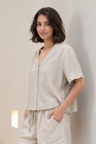 A woman with short brown hair wears the Olive Basics Bella Multi-way V-neck Shirt in Oatmeal, standing against a plain light-colored wall with her hands in her pockets.