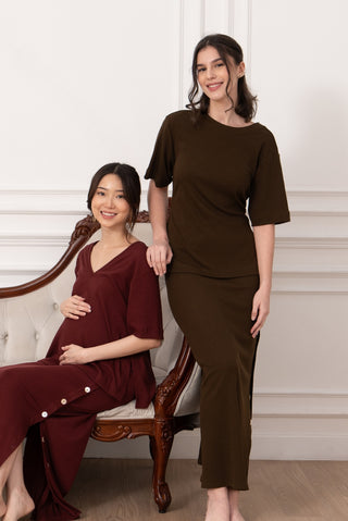 Two women pose indoors: one, who appears pregnant, sits on a vintage sofa in the Olive Basics Tabitha Reversible Multi-way Tee (TENCEL Cotton Rib) in burgundy; the other stands beside her in an oversized tee and dark brown skirt.