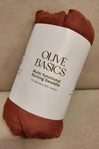 A rolled New York Pink Olive Basics Multi-functional Nursing Swaddle with a label, made of 70% bamboo and 30% cotton, is shown on a beige surface—ideal as a breathable baby blanket or nursing cover.
