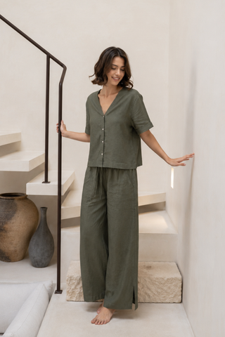 A woman stands barefoot indoors on a stone step, wearing Olive Basics’ Bella Multi-Pocket Drawstring Pants in khaki green and a loose button-up top, while holding the stair railing and touching the wall.
