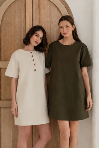 Two women stand indoors by a wooden arched door, each wearing the Olive Basics Joanna Reversible T-shirt Pocket Dress—one in cream with buttons, the other in olive green—both made from soft TENCEL cotton blend fabric.