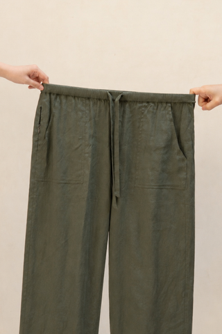 Two hands stretch the waistband of Olive Basics Bella Multi-Pocket Drawstring Pants in olive green TENCEL linen blend, featuring front pockets and a drawstring, set against a plain light background.