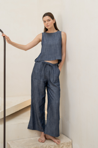 A woman stands barefoot on a stairway, wearing Olive Basics' sleeveless blue TENCEL™ linen blend top and Bella Multi-Pocket Drawstring Pants, with one hand in her pocket and the other resting on a railing.