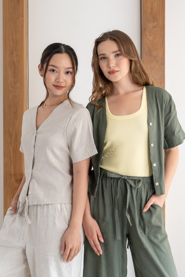 Two women stand side by side indoors, both dressed in casual, earth-toned outfits with relaxed silhouettes. One wears the Olive Basics Bella Multi-way V-neck Shirt, crafted from a soft linen blend and designed for nursing-friendly comfort. They face the camera with neutral expressions.