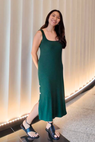 A woman wearing the Olive Basics Lydia Reversible Tank Dress stands indoors, smiling, in front of a curved, well-lit wall. She pairs the sleeveless green dress with black sandals.