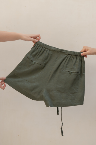 Two hands hold the waistband of Olive Basics’ Bella Multi-Pocket Drawstring Shorts, made from a dark green TENCEL linen blend with two buttoned back pockets, shown against a plain background.