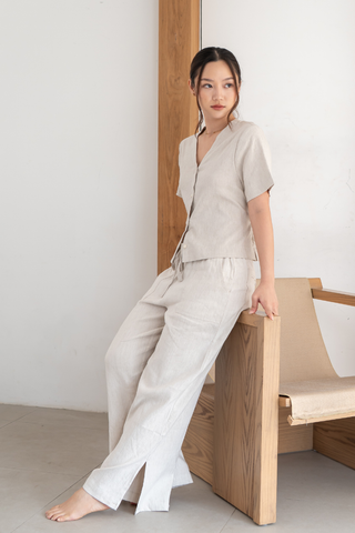 A woman, dressed in the Olive Basics Bella Multi-way V-neck Shirt—a light beige, linen-blend, nursing-friendly top—leans against a wooden chair in a minimalist room with white walls and a tiled floor.