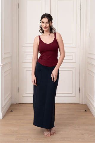 A woman stands barefoot on a wooden floor in front of white paneled double doors, wearing a sleeveless maroon top and the Olive Basics Michal Reversible Maxi Skirt with an adjustable waistband.