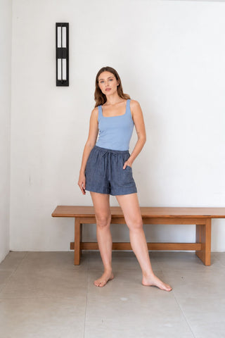 A woman wears the Olive Basics Esther Reversible Padded Tank in Sky Blue and blue shorts, standing barefoot indoors by a wooden bench and a white wall.