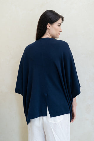 A woman with long dark hair faces away, wearing the Olive Basics Deborah Nursing Kimono Cardigan in navy blue and white pants against a light-colored wall.