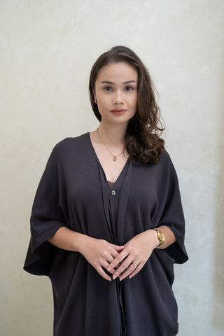 A woman with wavy brown hair wears the Olive Basics Deborah Nursing Kimono Cardigan, standing against a light textured wall and looking directly at the camera with a neutral expression.