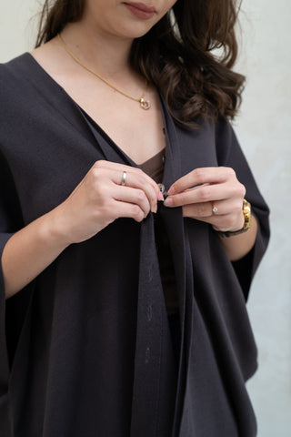 A woman buttons the Deborah Nursing Kimono Cardigan by Olive Basics over a brown top, accessorized with a gold necklace, ring, and wristwatch.