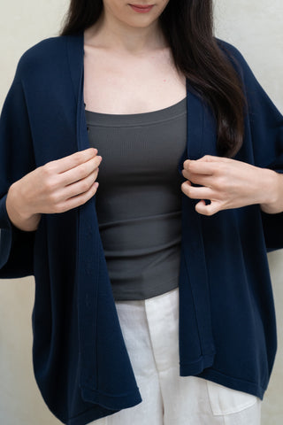 A woman stands against a light background, adjusting her Olive Basics Deborah Nursing Kimono Cardigan in dark blue, layered over a gray tank top and white pants.