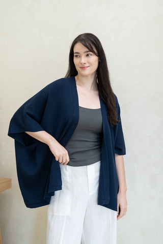 A woman with long dark hair wears the Olive Basics Deborah Nursing Kimono Cardigan in navy over a gray top and white pants, standing indoors against a light-colored wall.