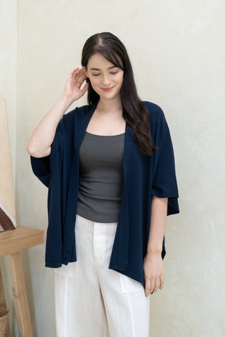 A woman with long dark hair stands indoors wearing the Olive Basics Deborah Nursing Kimono Cardigan in navy blue over a gray top and white pants, touching her hair with a neutral expression.
